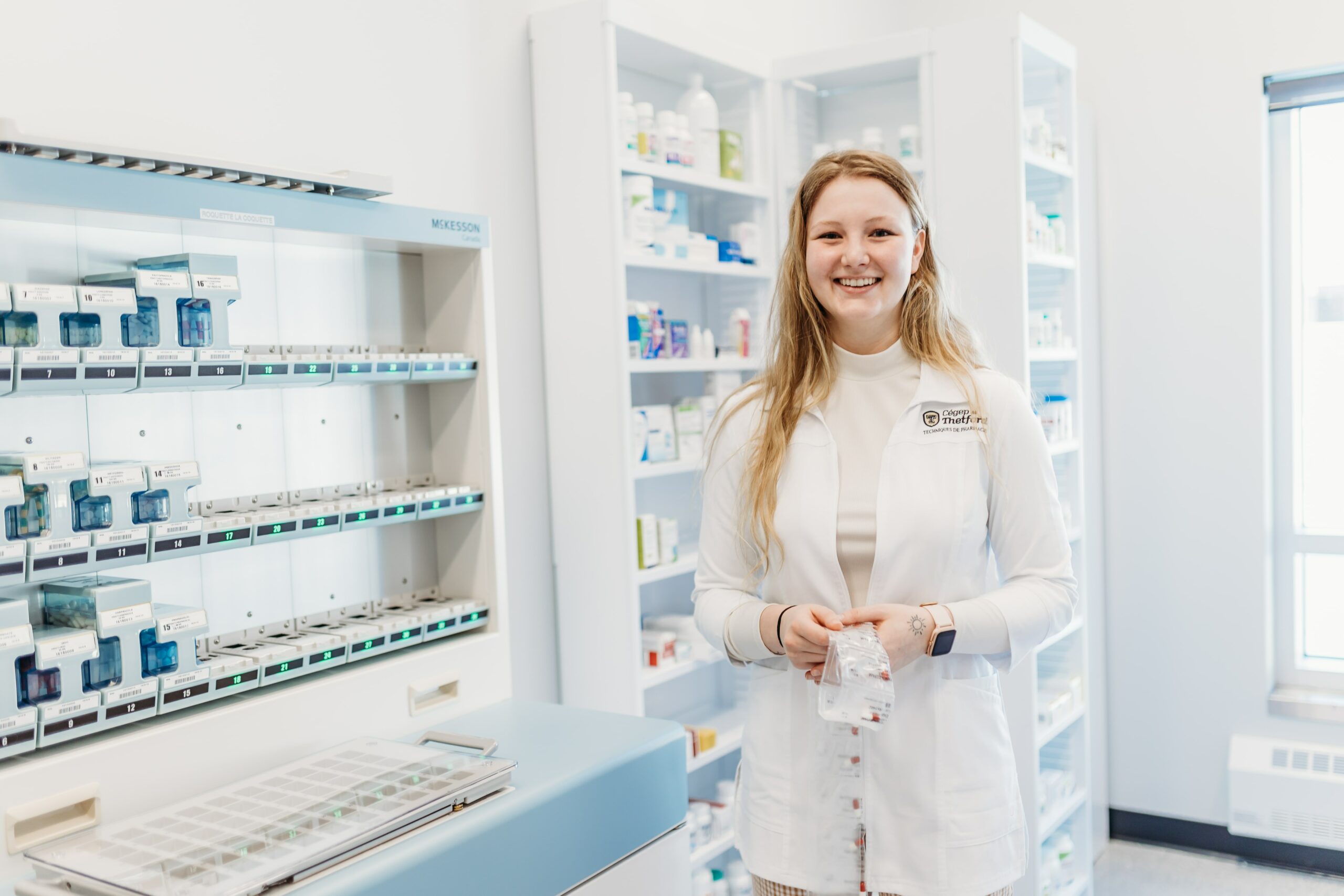 Une jeune femme sourit à la caméra dans un environnement de pharmacie.