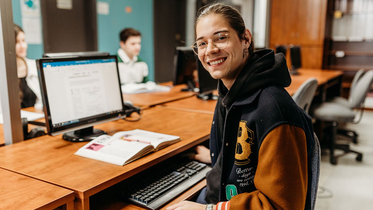 Un jeune homme souriant à la caméra devant un ordinateur.