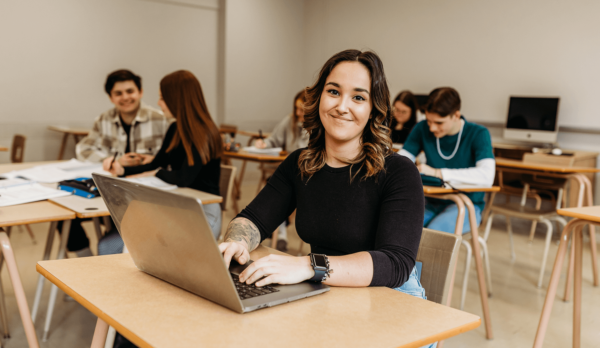 Une jeune femme souriant à la caméra dans une classe devant son ordinateur.