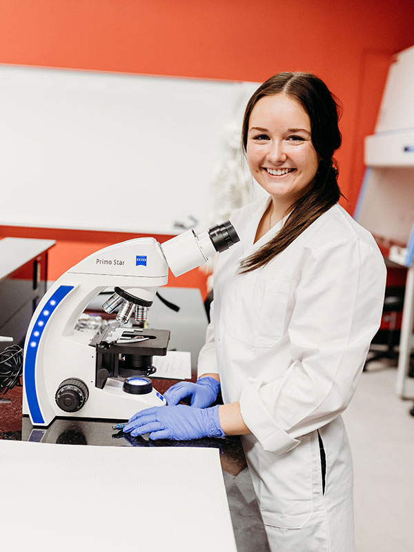Une étudiante souriant à la caméra au-dessus d'un microscope.