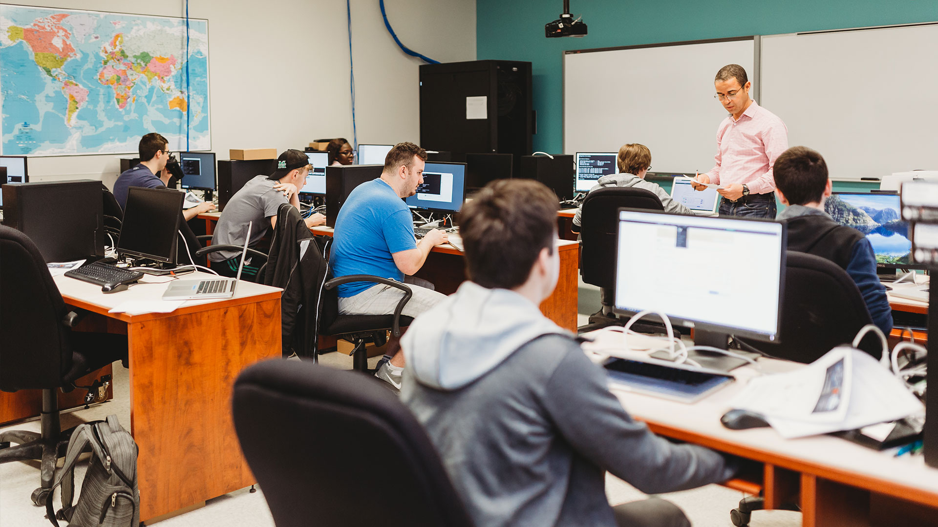 Photo d'une classe d'informatique collégiale avec un professeur debout donnant des consignes à des étudiants. Plusieurs ordinateurs et installations technologiques.