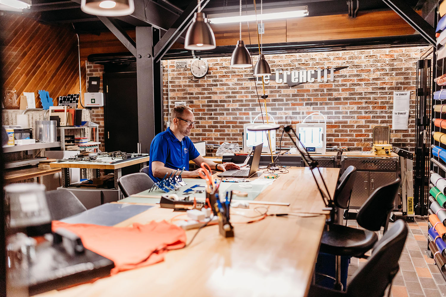 Photo d'un atelier créatif avec un homme travaillant sur un ordinateur portable. L'atelier possède un mur de brique, une longue table et plusieurs matériaux et échantillons au mur.