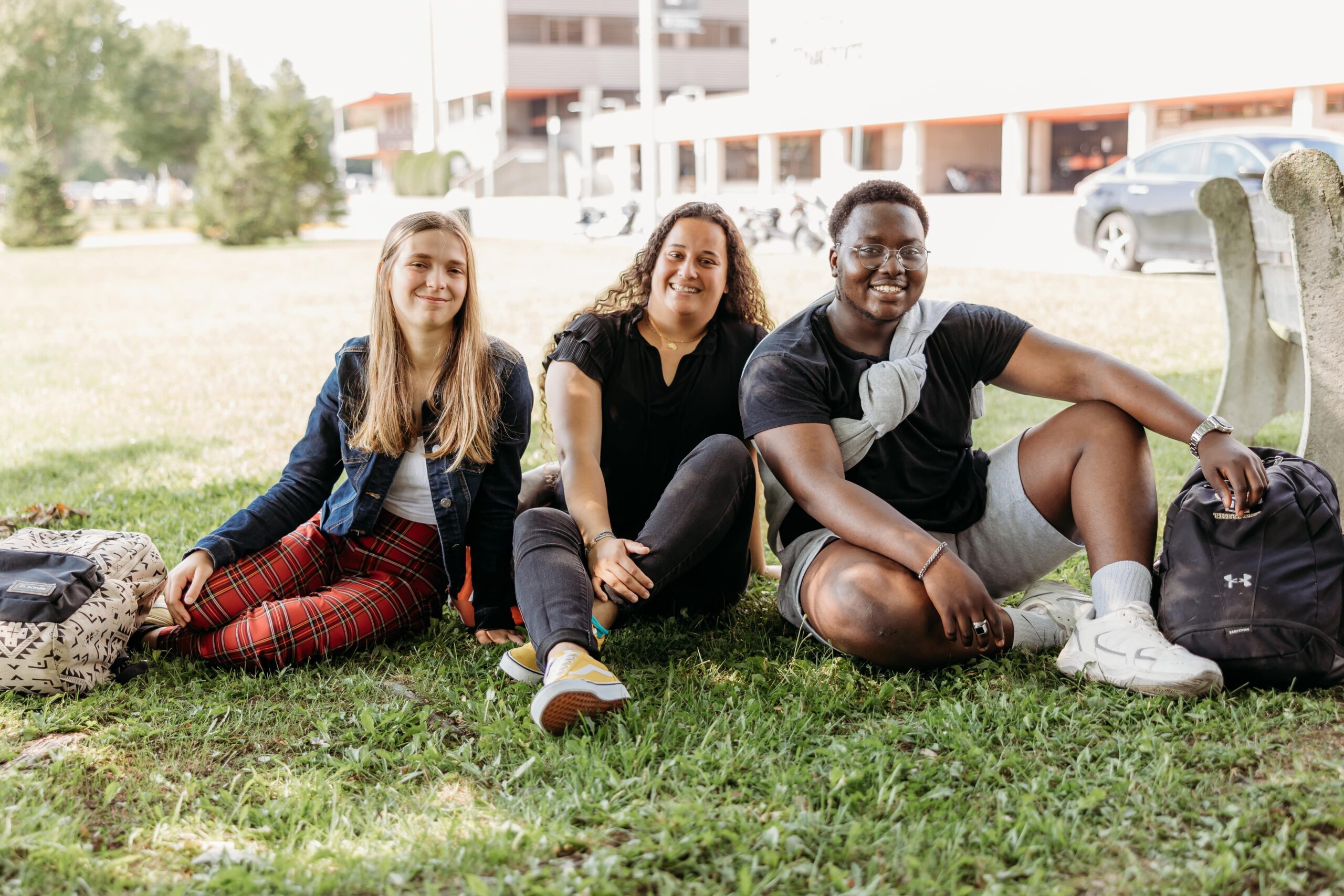 Trois étudiants souriants à la caméra assis sur le gazon à l'extérieur du Cégep de Thetford.