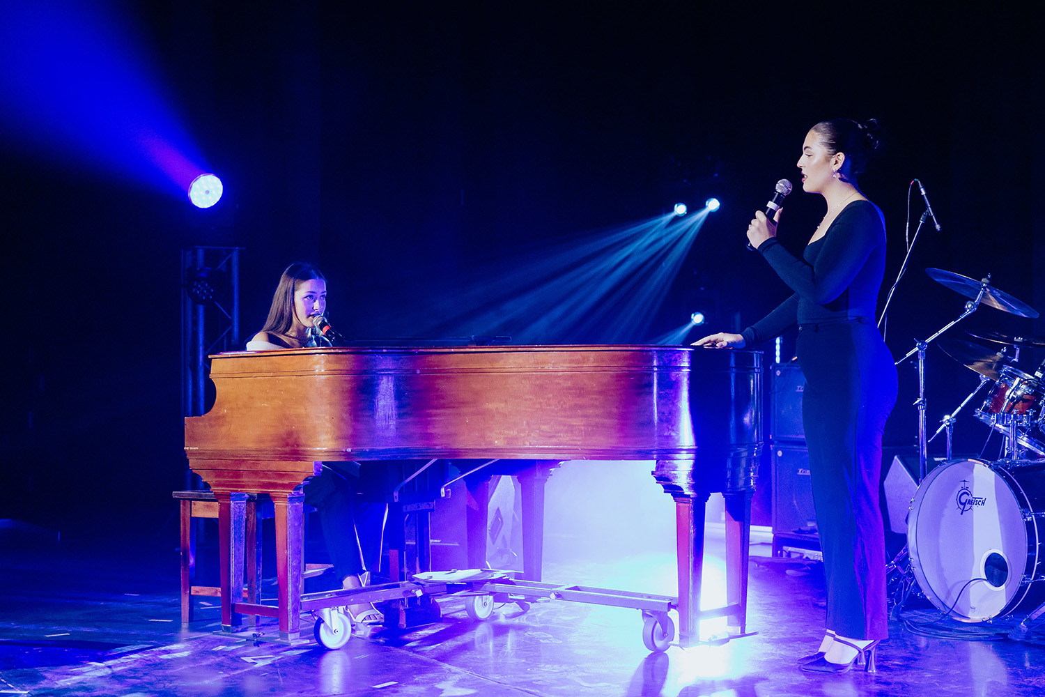 Des musiciens performent lors d'un spectacle. On retrouve une pianiste et une chanteuse debout, avec des lumières projecteurs sur la scène.