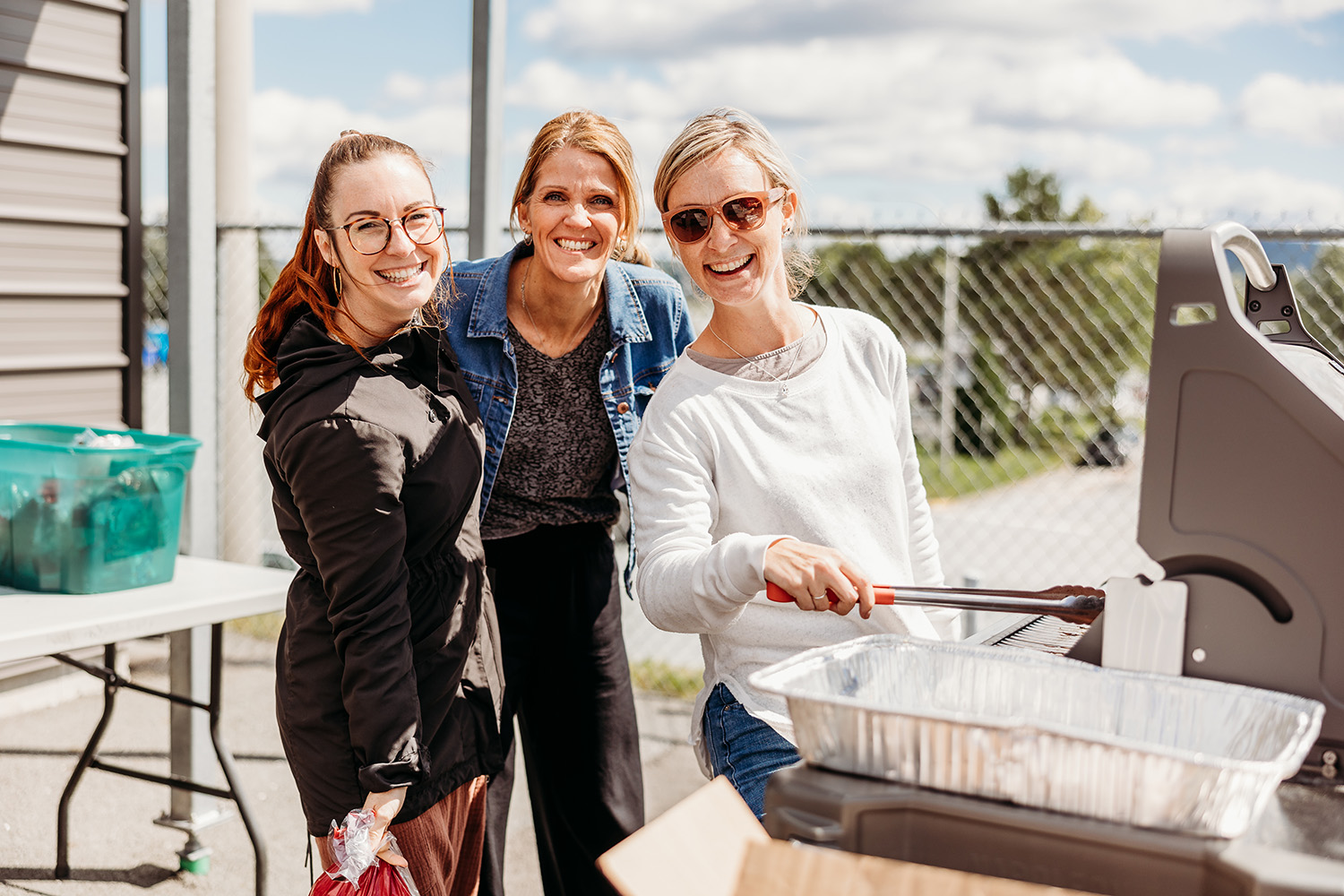 Trois jeunes femmes sourient à la caméra alors qu'elles font cuire de la nourriture sur un barbecue dans le cadre d'un événement social.