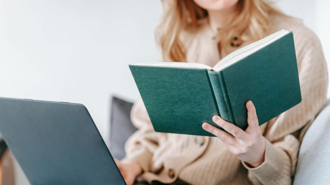 Gros plan qui présente une femme lisant un livre pendant qu'elle est sur son ordinateur portable.